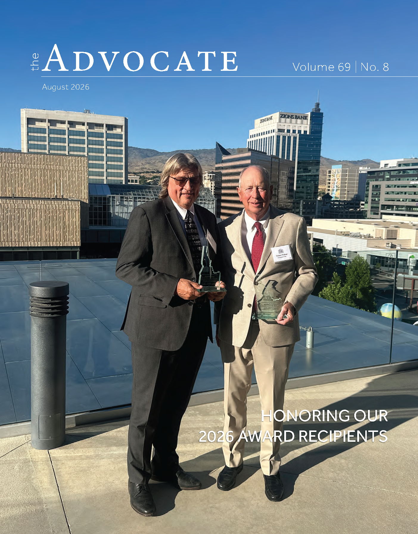 The Advocate magazine cover, Volume 69, No. 8, August 2026. Two men stand on a rooftop terrace overlooking downtown Boise, each holding a glass award shaped like the state of Idaho. The man on the left wears a dark suit with a patterned tie; the man on the right wears a tan sport coat with khaki pants and a red tie. Downtown Boise buildings, including the Zions Bank tower, and the foothills are visible in the background under a clear blue sky. White text at the bottom right reads 'Honoring Our 2026 Award Recipients.
