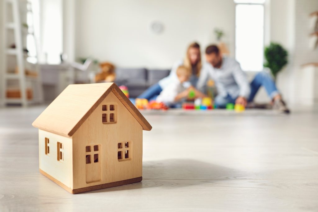 Close-up of miniature detached house on floor at home with happy family playing with little child in background.