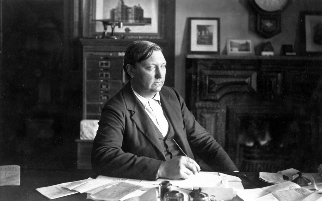 Man at desk in late 1800's in suit with papers at desk.