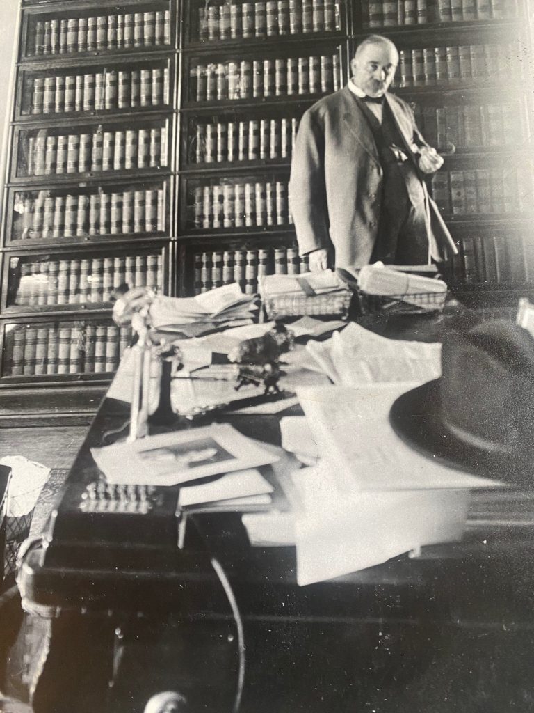 Man in 1900's standing behind desk with many papers in office.