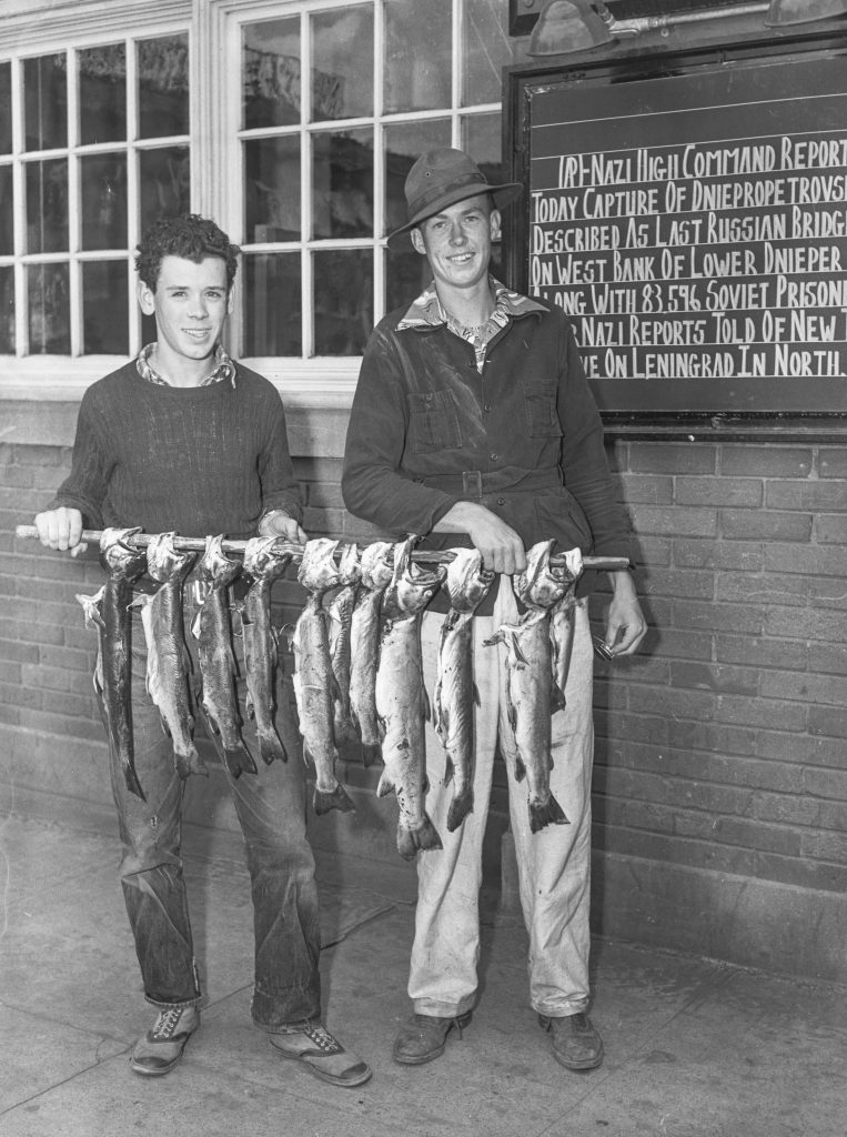 Two men stand with fish in front of the Statesmen office which has updates from the War.