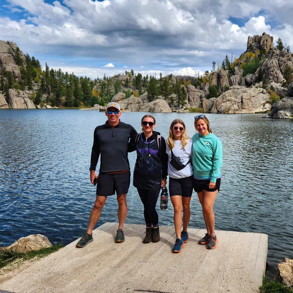 maureen and husband and 2 daughters post in front of mountain lake,