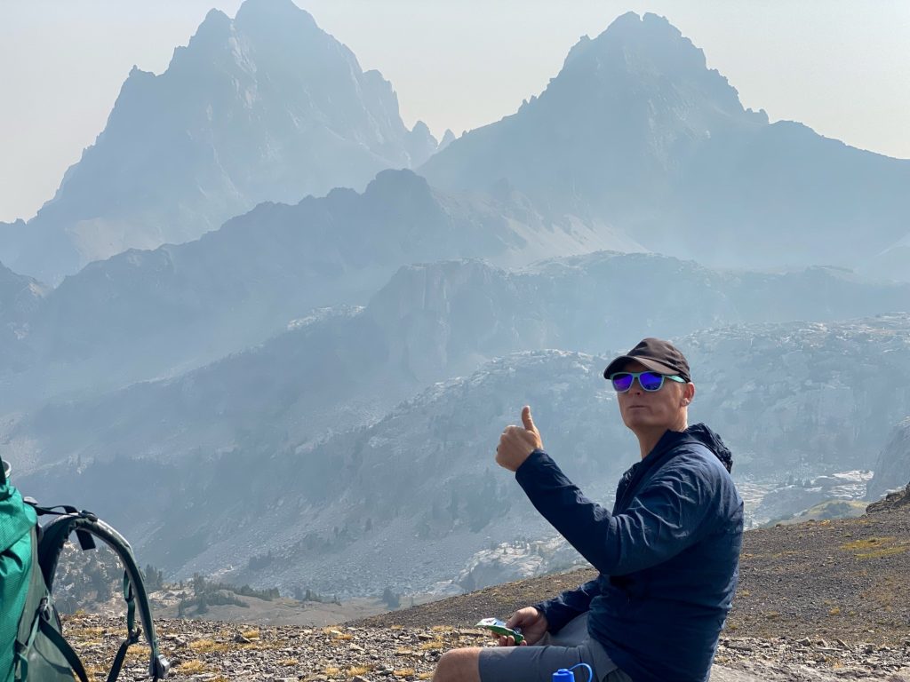 Man backpacking with thumbs up in front of mountain range.